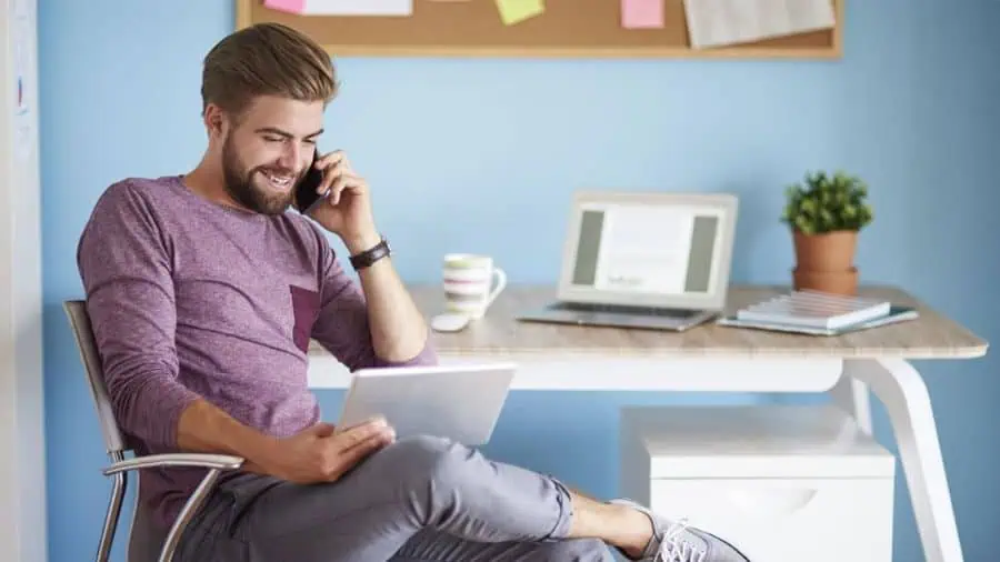 A man sitting in a chair talking on a phone and checking his flexitime balance on a tablet